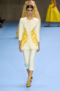Model walking the runway in a white structured coat with yellow floral embroidery along the front edges, paired with cropped white pants.
