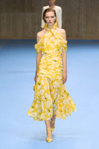 Model walks a bright yellow floral halter-neck dress with off-shoulder sleeves on a blue runway runway.