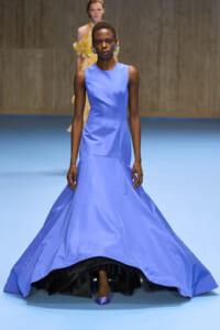 Model walks the runway in a sleeveless lavender-blue gown with a full, floor-sweeping skirt on a blue runway surface.