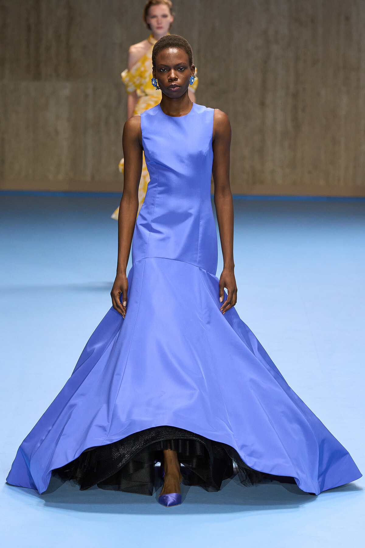 Model walks the runway in a sleeveless lavender-blue gown with a full, floor-sweeping skirt on a blue runway surface.