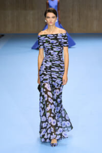 Model wearing an off-the-shoulder, floor-length black gown with blue floral print walking a blue runway.