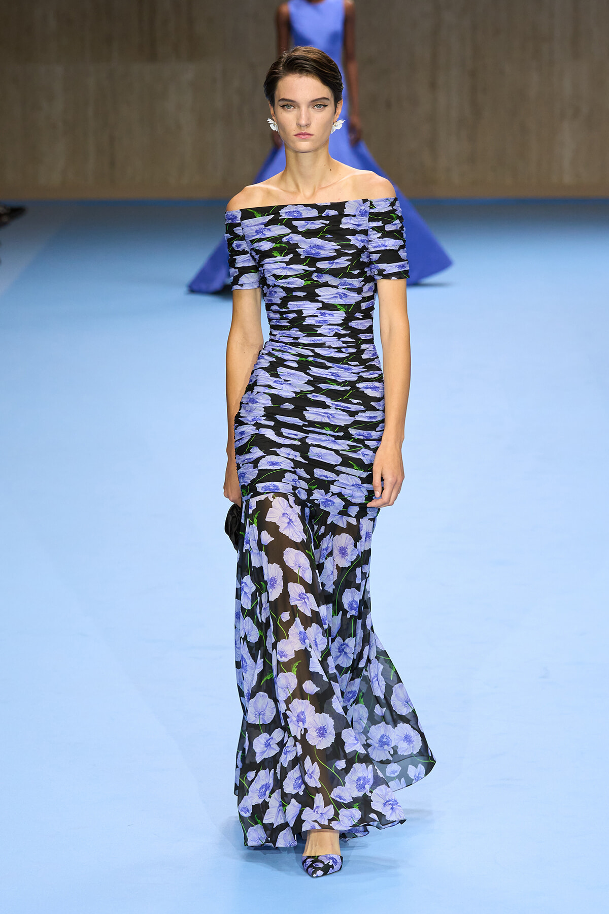 Model wearing an off-the-shoulder, floor-length black gown with blue floral print walking a blue runway.