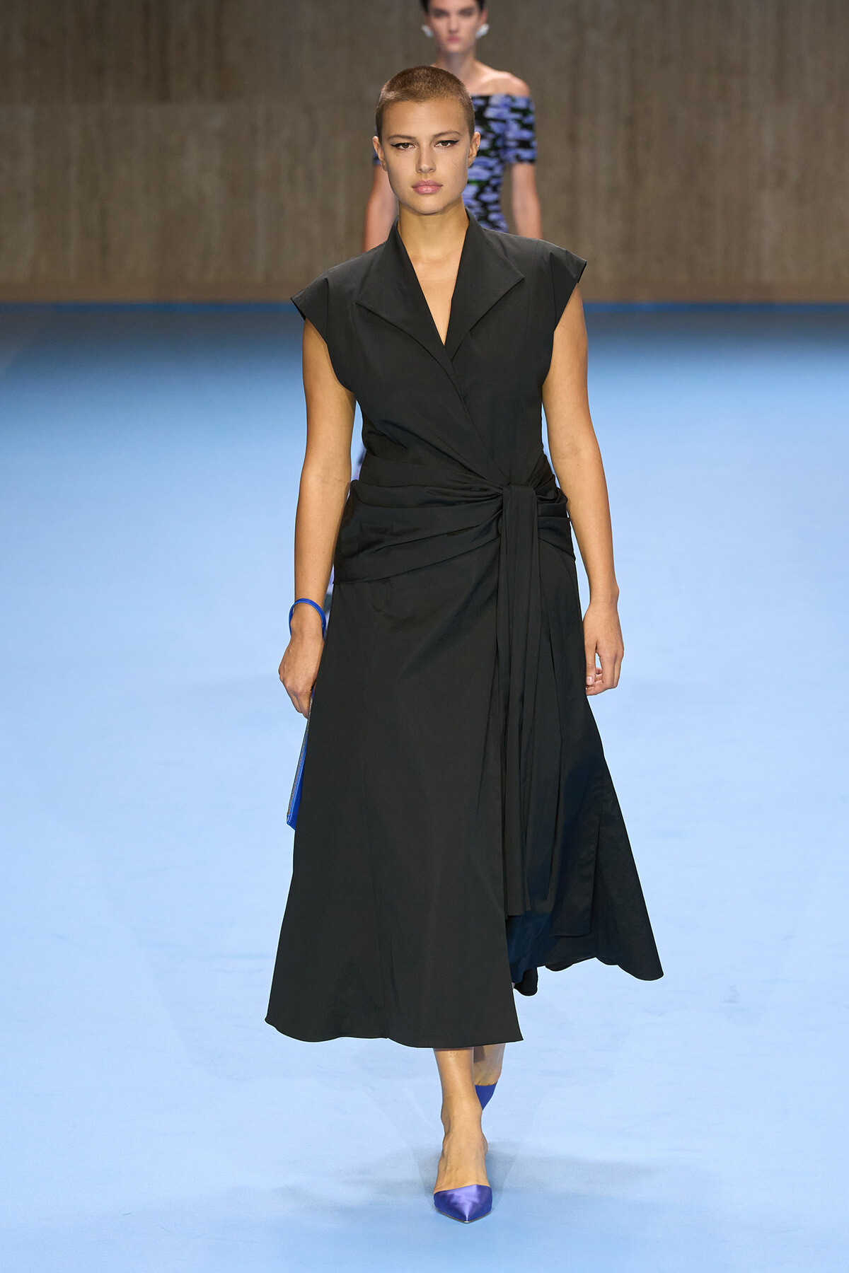 Model walking the runway in a black sleeveless wrap dress with a tied waist on a blue catwalk, wearing blue heels.