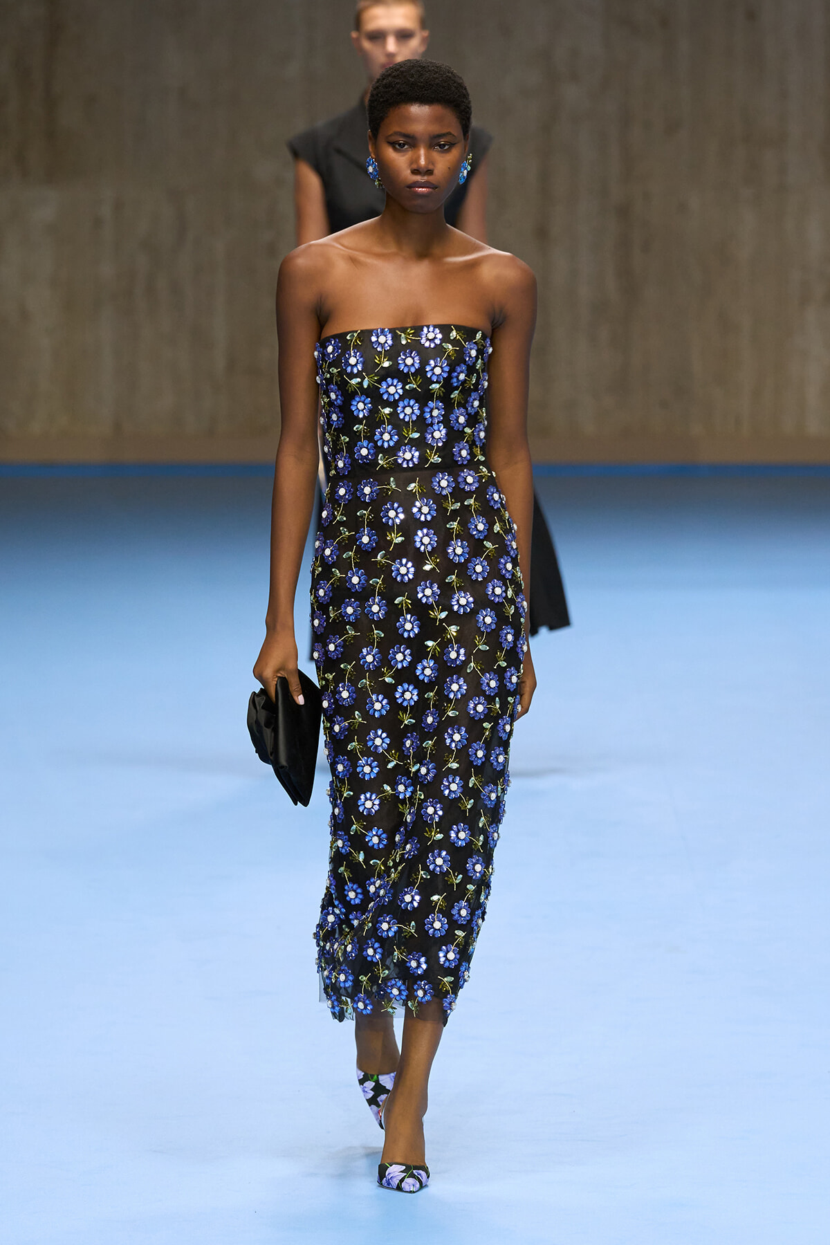 Model walks a fashion show wearing a strapless black midi dress with blue floral print and matching blue earrings.