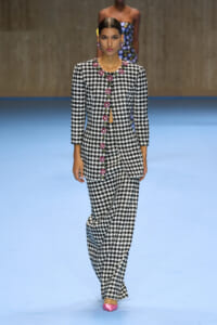Model walking a runway in a black-and-white houndstooth suit with pink floral button trim and matching wide-leg pants, wearing pink pointed-toe heels.