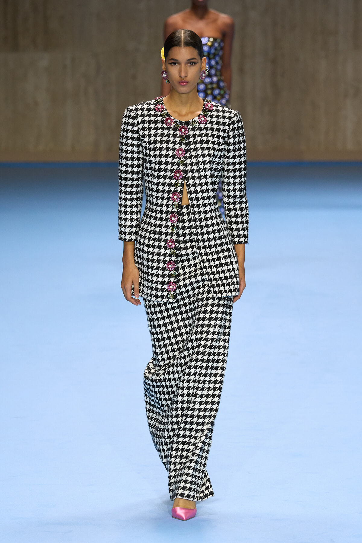 Model walking a runway in a black-and-white houndstooth suit with pink floral button trim and matching wide-leg pants, wearing pink pointed-toe heels.