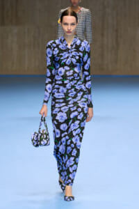 Model walks the runway in a blue floral-print matching set (top and pants) with a small matching handbag and flats.