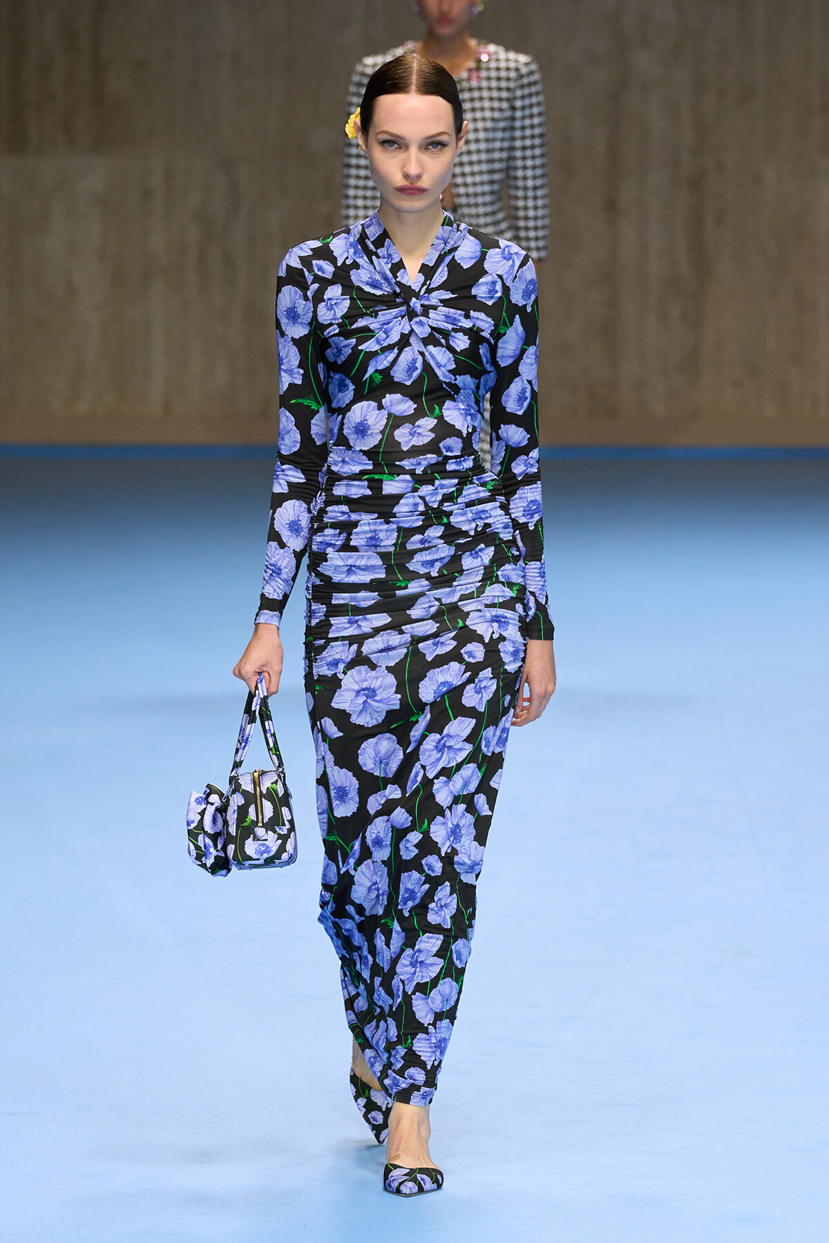 Model walks the runway in a blue floral-print matching set (top and pants) with a small matching handbag and flats.