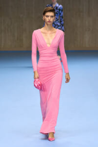 Model wearing a pink, form-fitting gown with a plunging V-neckline and ruching at the waist, walking a fashion show while holding a matching pink clutch.