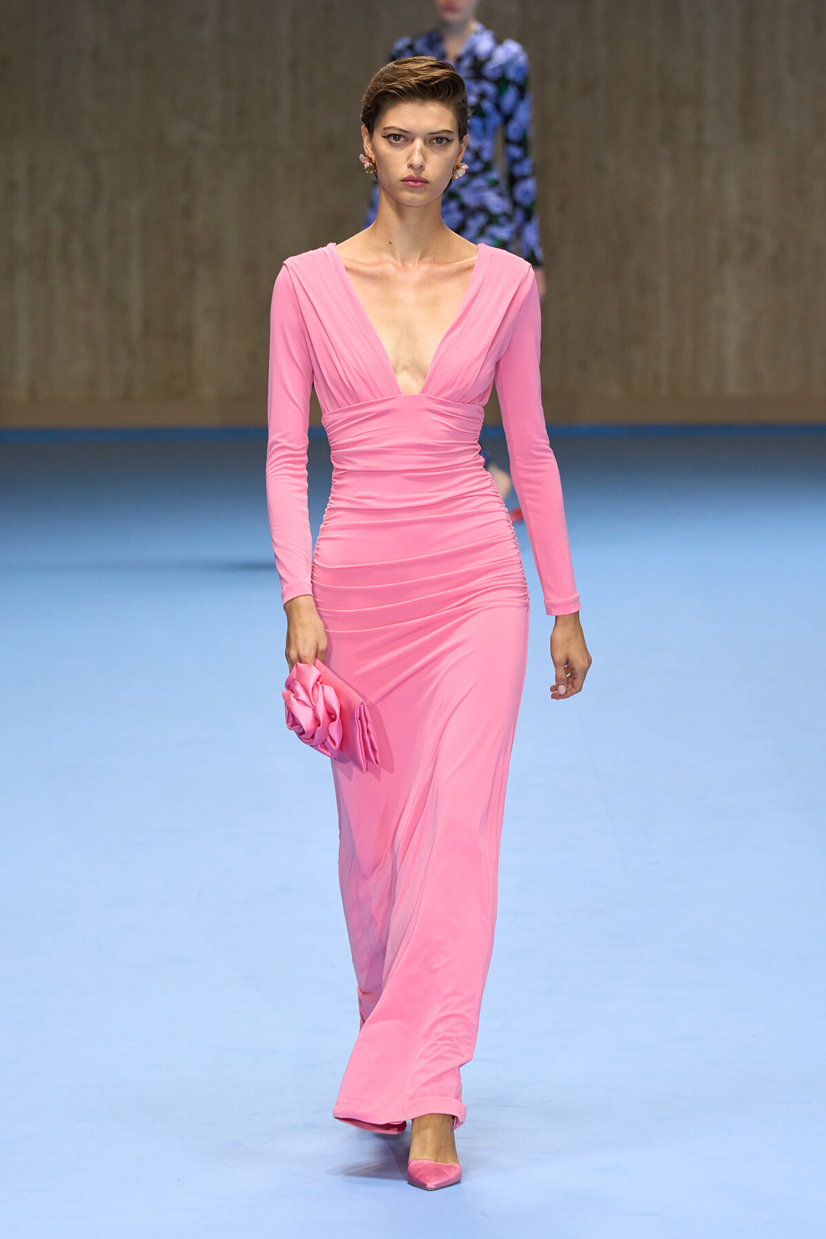 Model wearing a pink, form-fitting gown with a plunging V-neckline and ruching at the waist, walking a fashion show while holding a matching pink clutch.