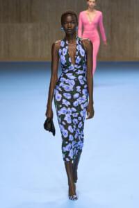 Model walking a fashion show in a blue-flowered black halter-neck dress, carrying a small black clutch.