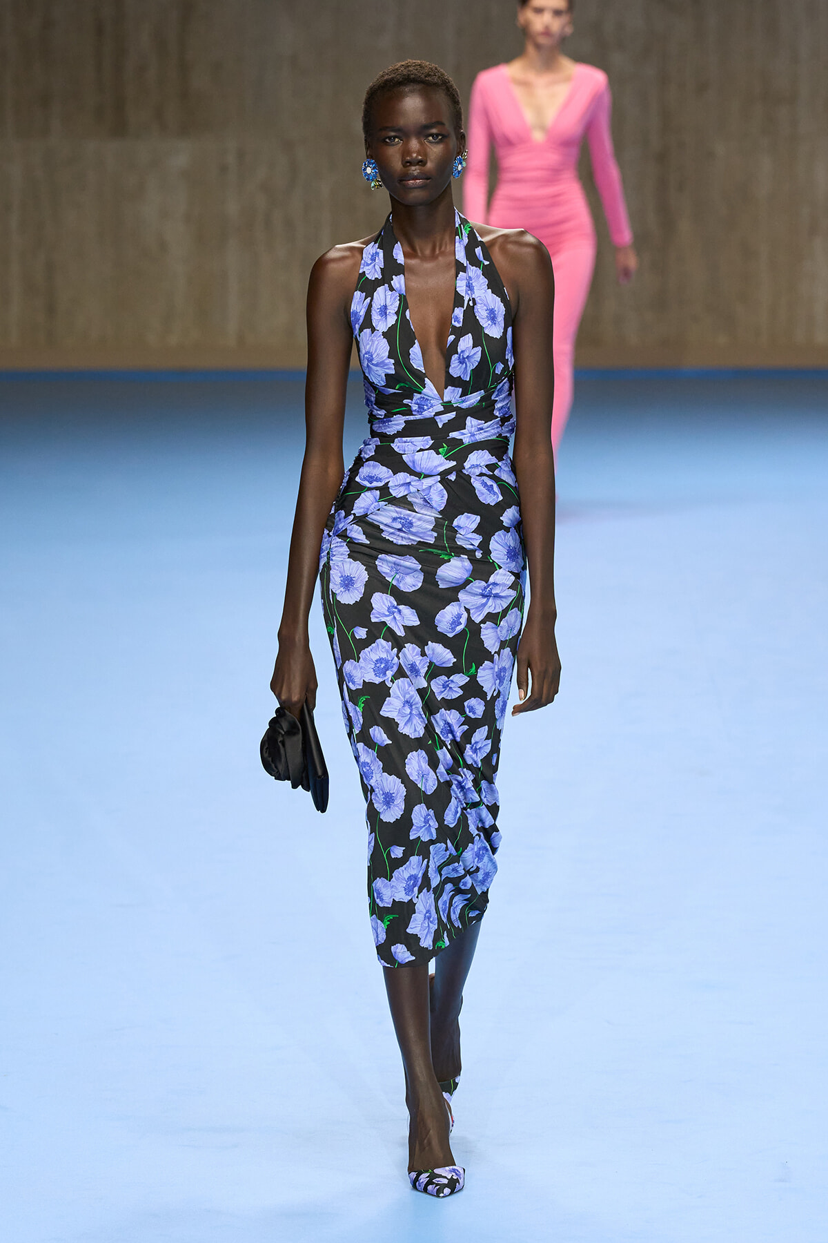 Model walking a fashion show in a blue-flowered black halter-neck dress, carrying a small black clutch.