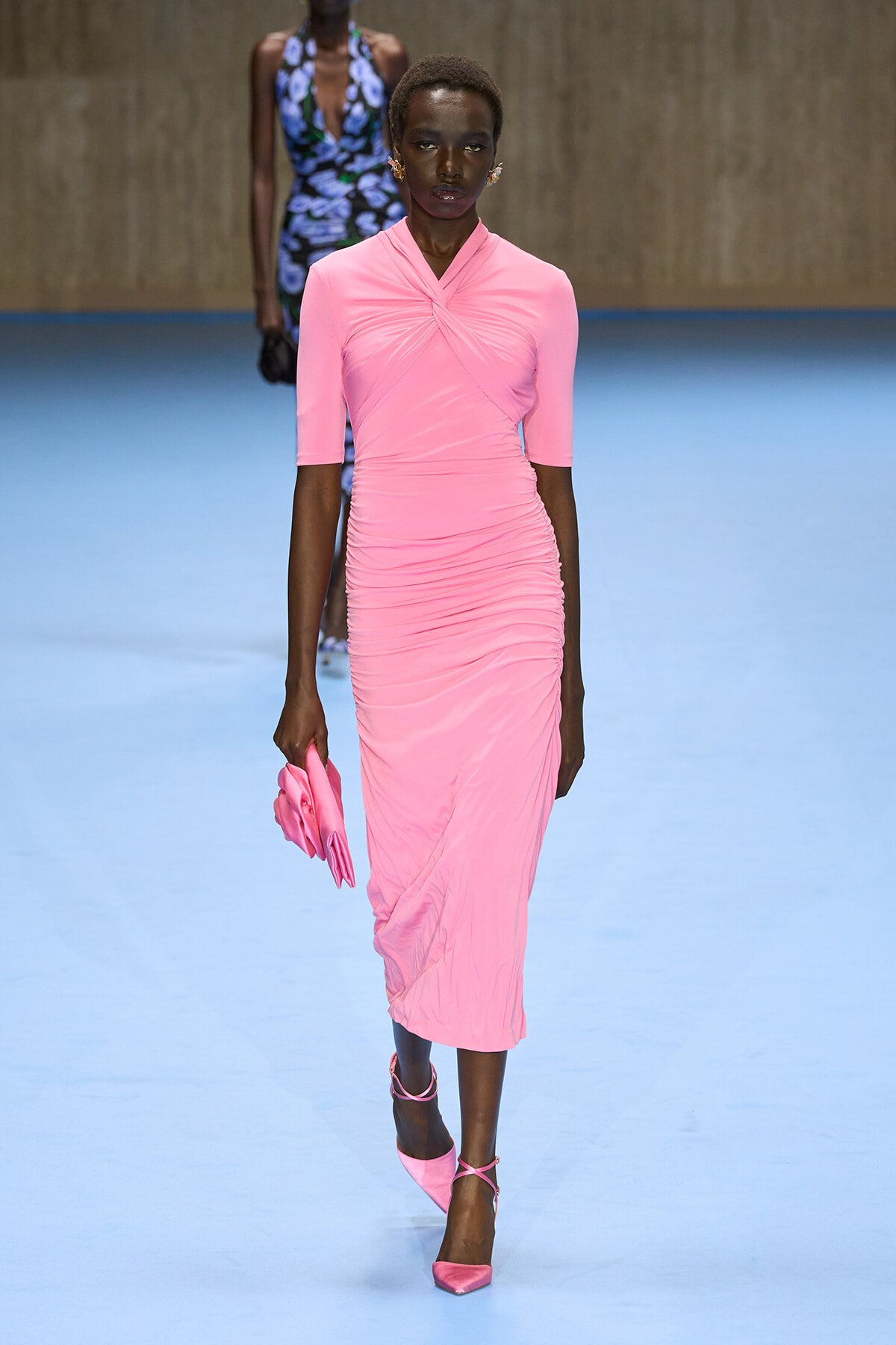 Model walking a runway in a knee-length pink ruched dress with a twist-front neckline, holding a pink clutch and wearing matching pink strappy heels on a light blue catwalk.