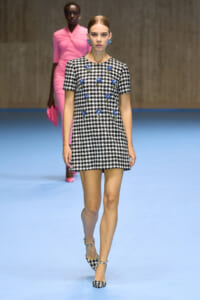 Model on a blue runway wearing a black-and-white houndstooth mini dress with blue floral embellishments, matching heels, and blue earrings.