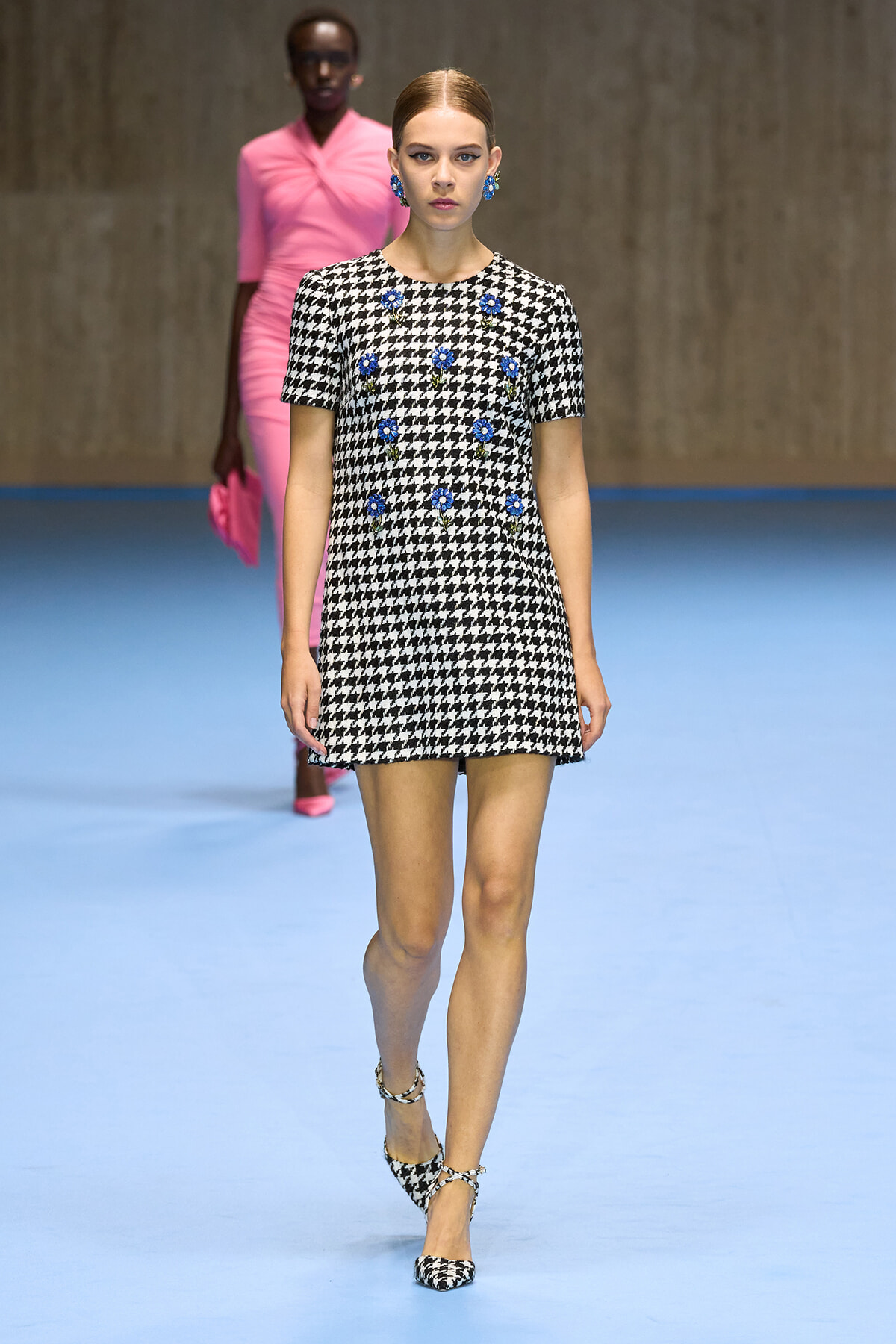 Model on a blue runway wearing a black-and-white houndstooth mini dress with blue floral embellishments, matching heels, and blue earrings.