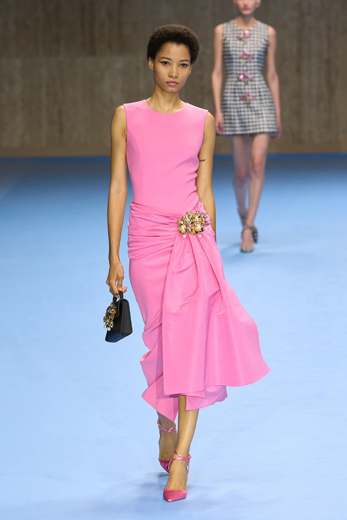 Runway model in a bright pink sleeveless dress with a jeweled floral cluster at the hip, carrying a small black clutch on a blue catwalk.