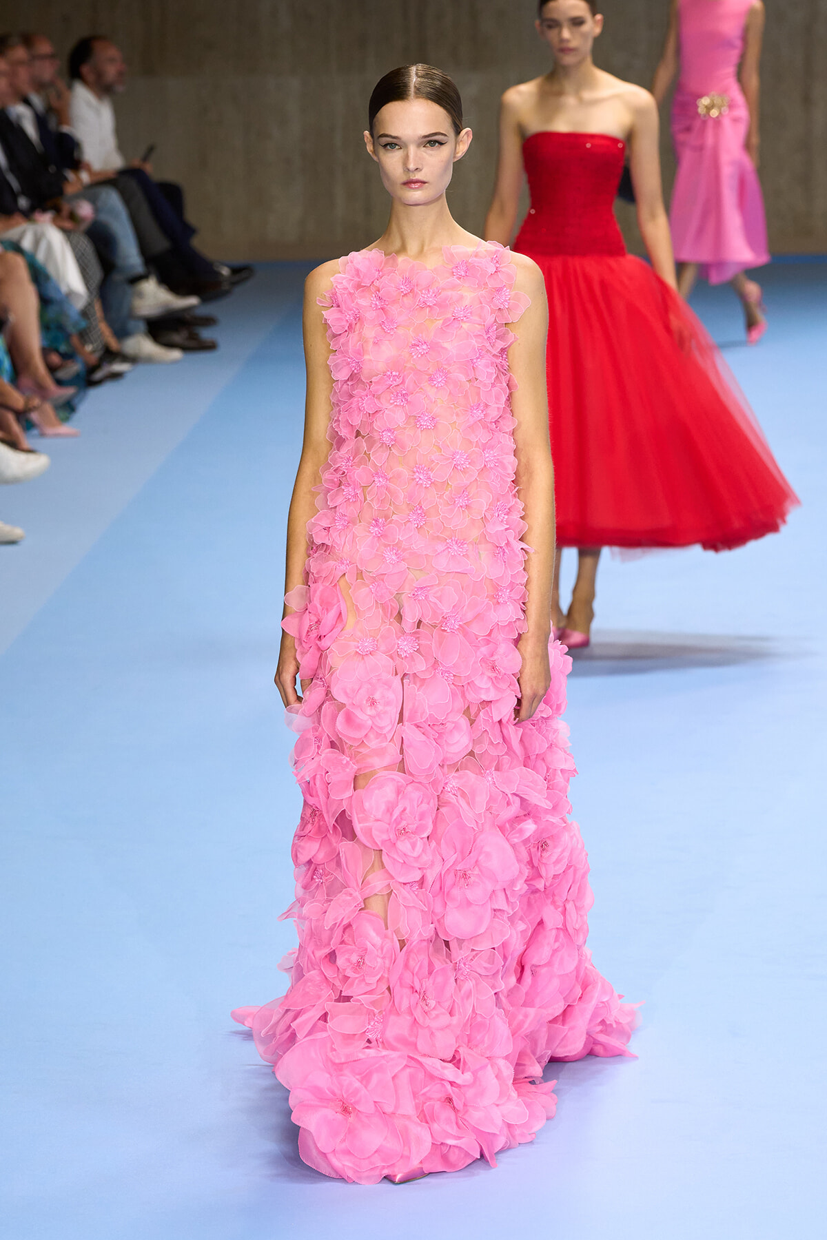 Model walking the runway in a pink, 3D-flower gown at a fashion show on a blue catwalk (audience visible to the left).