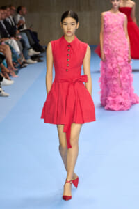 Model walking a fashion show in a sleeveless red dress with a belt and button placket on a blue runway.