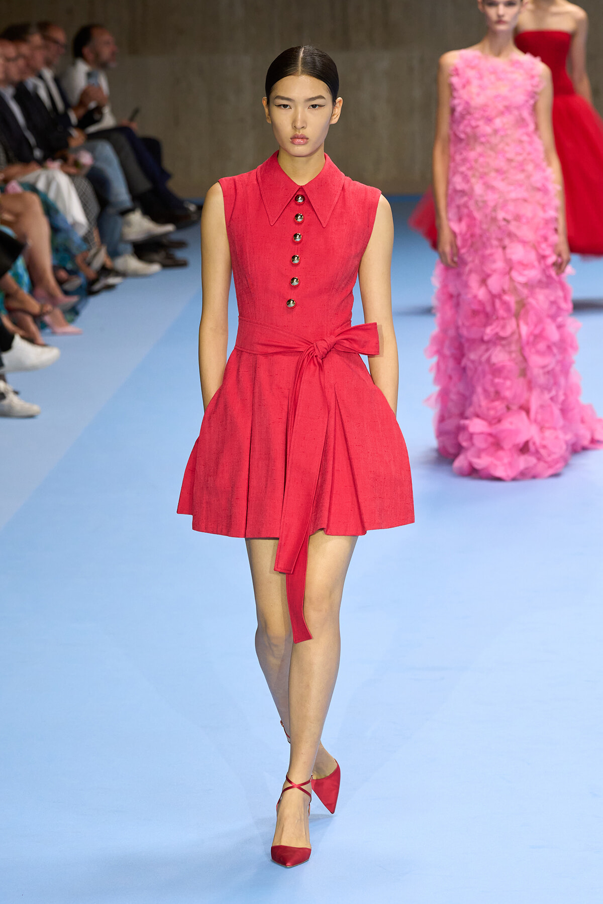 Model walking a fashion show in a sleeveless red dress with a belt and button placket on a blue runway.