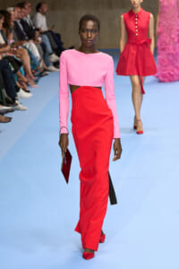 Model struts the runway in a pink cropped long-sleeve top and a high-waisted red pencil skirt, clutch in hand, audience in the background.