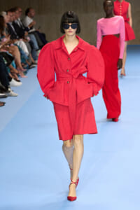 Model walks the runway in a red belted ensemble with puff sleeves and matching skirt, wearing dark sunglasses.