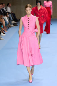 Model walks the runway in a pink sleeveless midi dress with a buttoned bodice and a waist bow, blue catwalk backdrop.