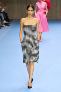 Model walking down a blue runway in a strapless black-and-white houndstooth midi dress with pockets, wearing black pointed heels.