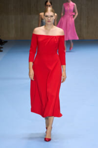 Model walking the runway in a red off-the-shoulder midi dress with three-quarter sleeves and matching red heels, on a light blue catwalk.