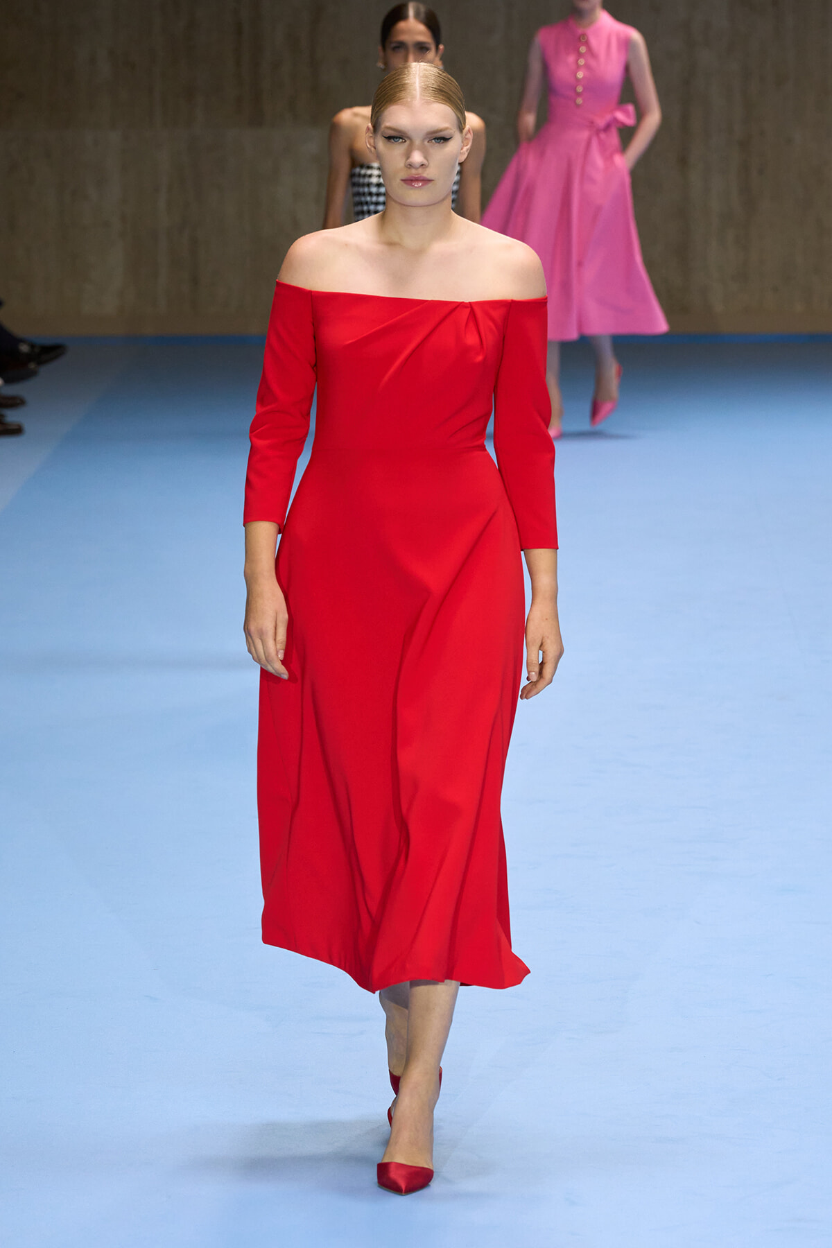 Model walking the runway in a red off-the-shoulder midi dress with three-quarter sleeves and matching red heels, on a light blue catwalk.