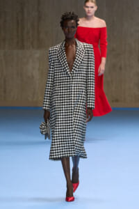 Model walks the runway in a black-and-white houndstooth coat with a deep V, carrying a matching houndstooth clutch and red pointed-toe heels.