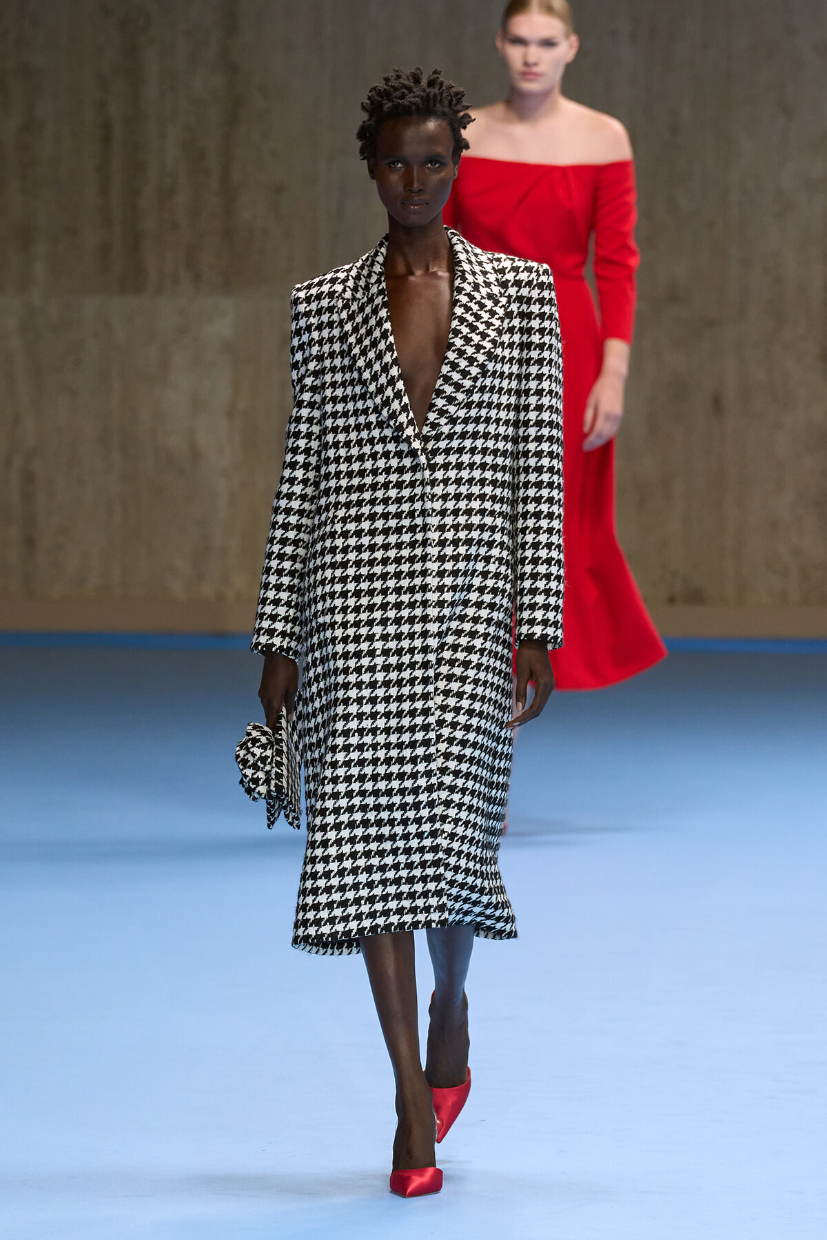 Model walks the runway in a black-and-white houndstooth coat with a deep V, carrying a matching houndstooth clutch and red pointed-toe heels.