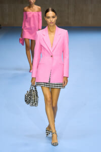 Model walks a runway in a bright pink blazer over a black-and-white houndstooth mini skirt, carrying a matching handbag.