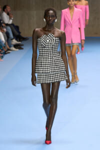 Runway model wears a strapless black-and-white houndstooth mini dress with red pointed heels on a blue catwalk.