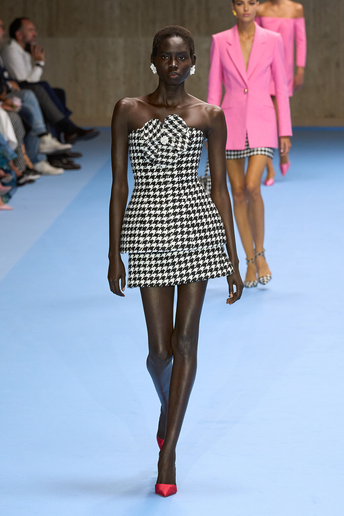 Runway model wears a strapless black-and-white houndstooth mini dress with red pointed heels on a blue catwalk.