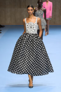 Model walks the runway in a sleeveless white top with black polka dots and a large fabric flower at the bodice, paired with a full black skirt with white polka dots on a blue catwalk.