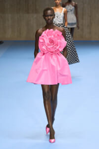 Model walks a blue runway wearing a bright pink strapless mini dress with a large fabric rose at the bodice and matching pink heels.