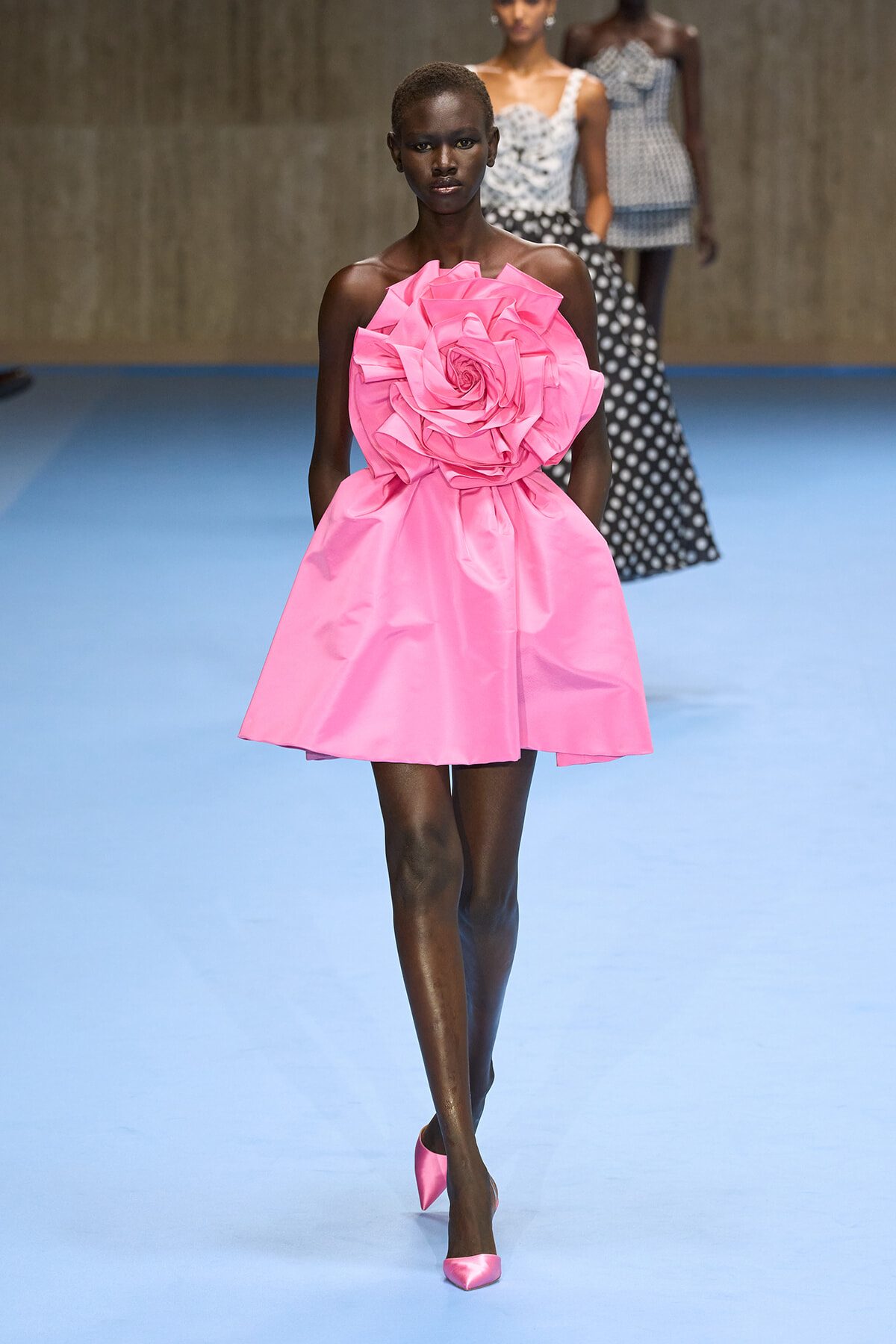 Model walks a blue runway wearing a bright pink strapless mini dress with a large fabric rose at the bodice and matching pink heels.