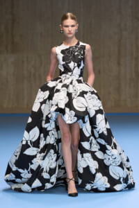 Model on a runway wearing a sleeveless black-and-white floral ball gown with a dramatic voluminous skirt and black heels.