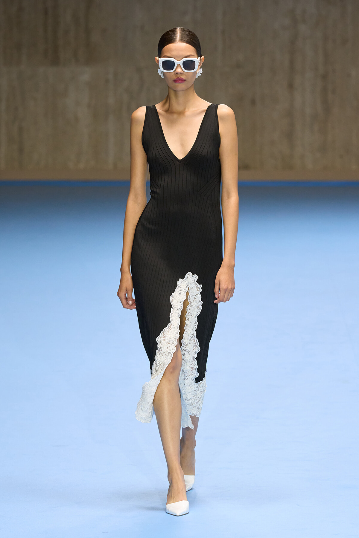 Model walking a runway in a black sleeveless knit dress with a white lace-trimmed front slit, paired with white sunglasses and white heels on a blue catwalk.