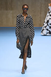 Model in black-and-white polka-dot blouse and high-waisted polka-dot skirt with lace hem, wearing oversized sunglasses on a blue fashion runway.