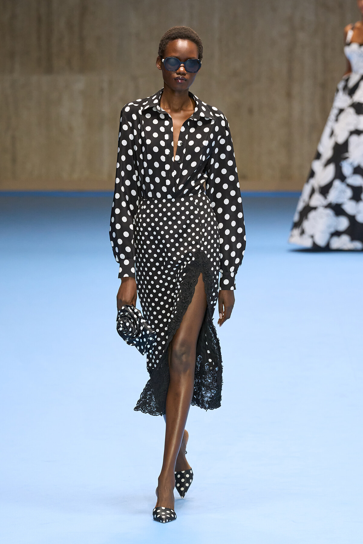 Model in black-and-white polka-dot blouse and high-waisted polka-dot skirt with lace hem, wearing oversized sunglasses on a blue fashion runway.