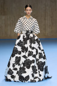 Model wearing a white blouse with large black polka dots and a voluminous black-and-white floral ball gown on a runway, belt cinching the waist.