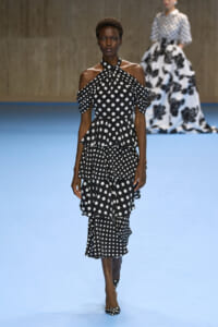 Model walks the runway in a black polka-dot halter-neck dress with off-shoulder sleeves and a three-tier ruffled skirt.