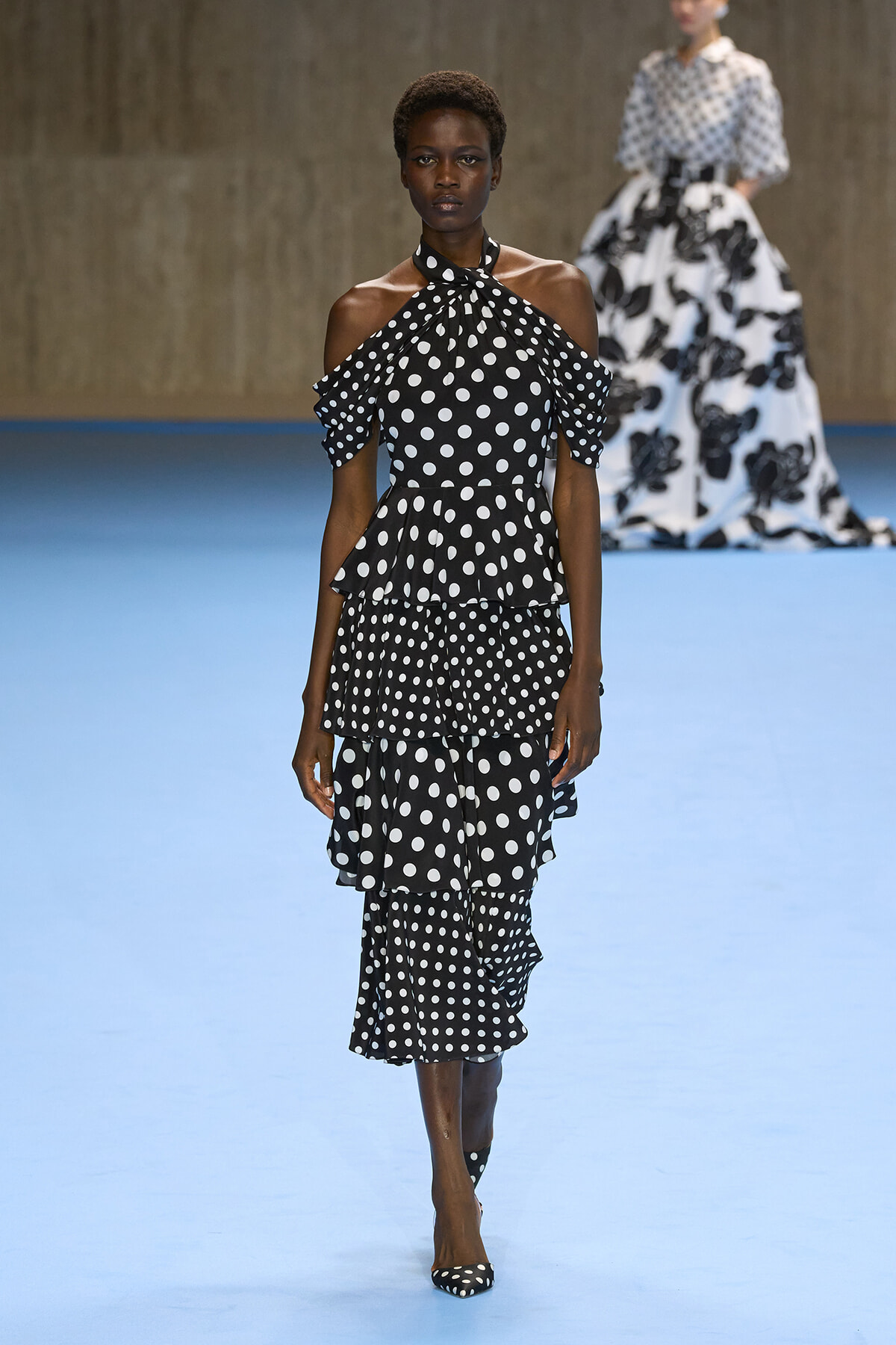 Model walks the runway in a black polka-dot halter-neck dress with off-shoulder sleeves and a three-tier ruffled skirt.
