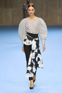 Model on a blue runway wears a white polka-dot blouse with oversized puff sleeves and a black-and-white floral wrap skirt over black pants paired with heels.