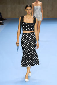 Runway model wearing a black-and-white polka-dot crop top with tied straps and a matching high-waisted midi skirt, carrying a light-blue clutch.