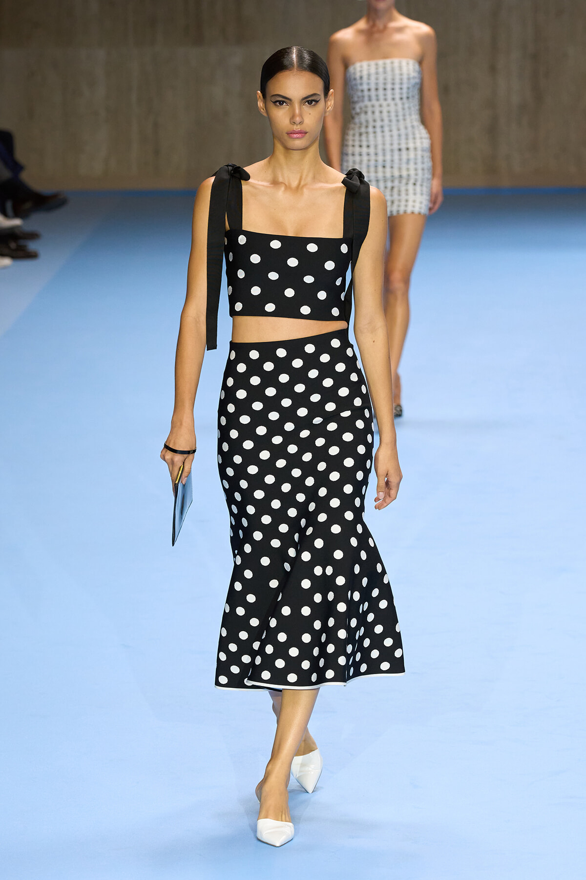 Runway model wearing a black-and-white polka-dot crop top with tied straps and a matching high-waisted midi skirt, carrying a light-blue clutch.