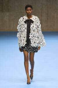 Model walking runway in a white and black polka-dot ensemble with a ruffled coat, black bow tie, and matching polka-dot heels.