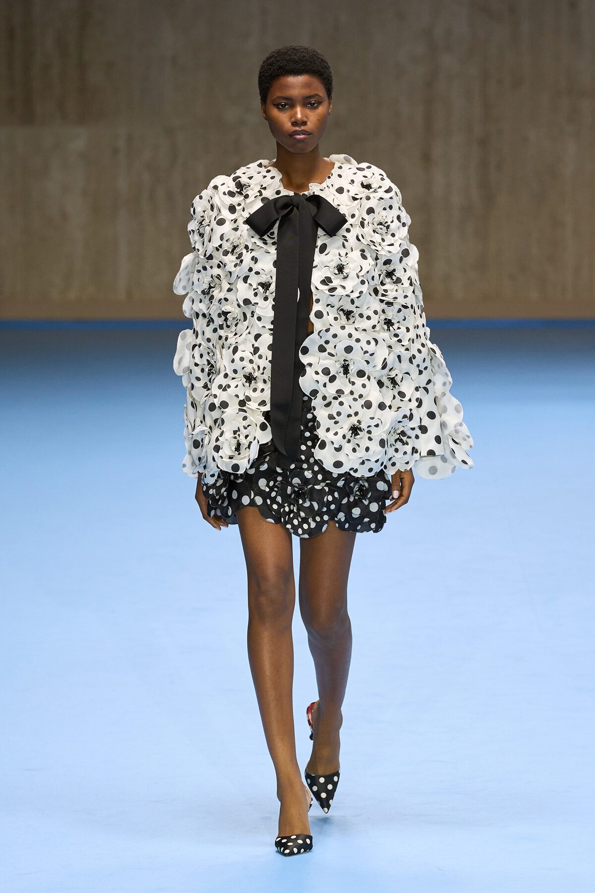 Model walking runway in a white and black polka-dot ensemble with a ruffled coat, black bow tie, and matching polka-dot heels.
