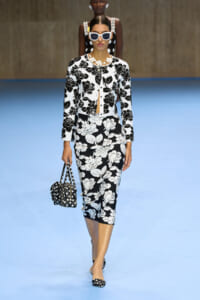 Model walking runway in black-and-white floral suit with oversized white sunglasses and matching polka dot bag and shoes, blue floor backdrop.